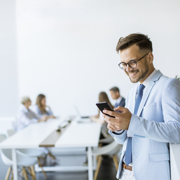 Happy young man using his mobile phone and smiling while his colleagues working in the background