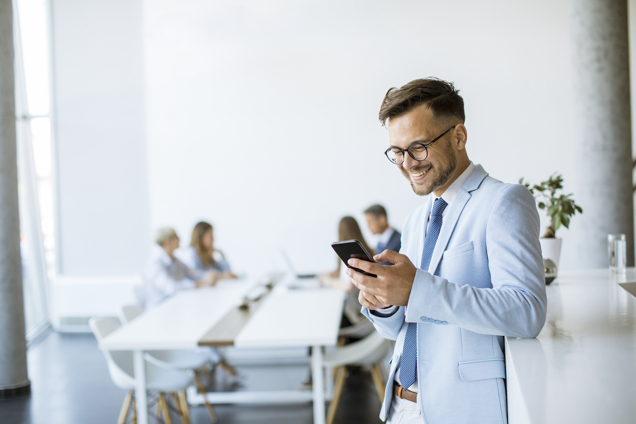 Happy young man using his mobile phone and smiling while his colleagues working in the background