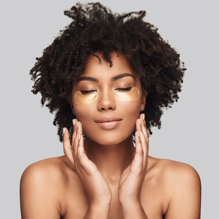 kin care. Attractive young African woman applying medical eye patch and smiling while standing against grey background