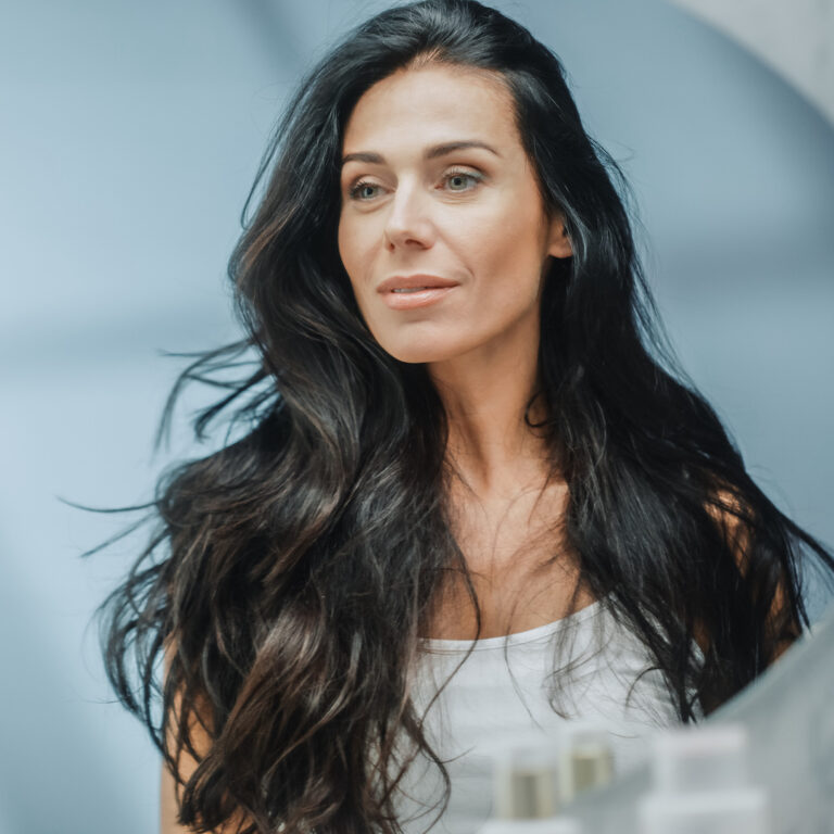 Beautiful Caucasian Middle Aged Woman Looks into Bathroom Mirror, Enjoys Her Looks. Concept for Happiness, Wellbeing, Natural Beauty and Organic Skin Care Products