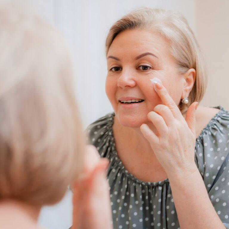 Happy healthy Mature woman at mirror apply anti aging moisturizing cosmetic cream on face, smiling middle-aged lady soft clean skin care and beauty