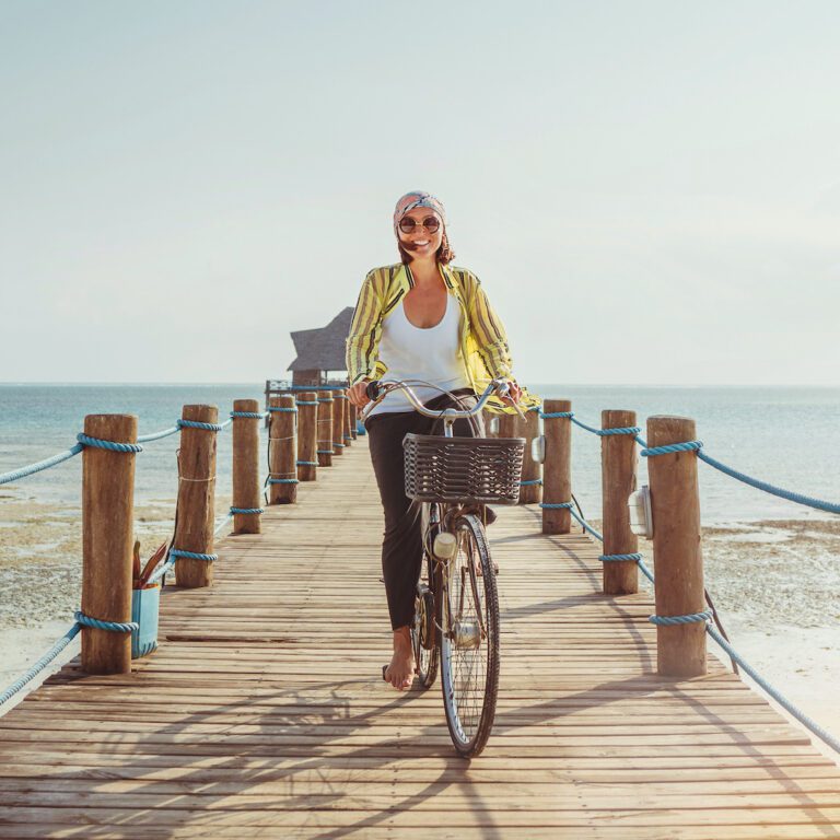 Portrait of a happy smiling woman dressed in light summer clothes and sunglasses riding a bicycle on the wooden sea pier and looking at camera. Careless vacation in tropical countries concept image